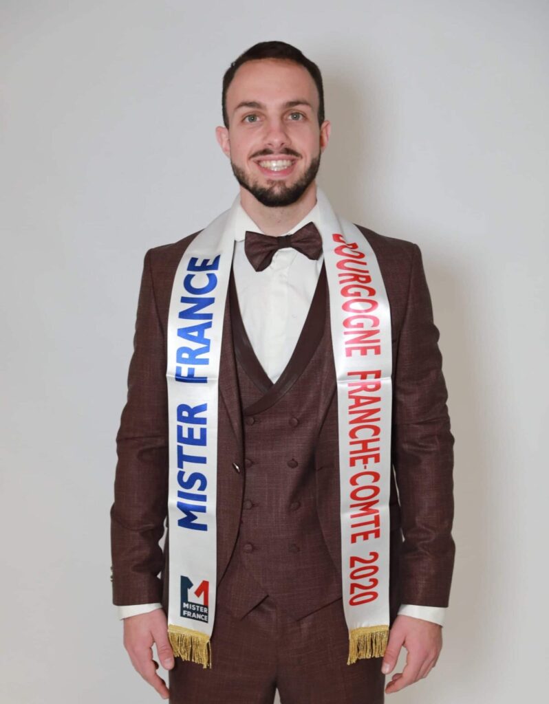 Théo SAUVREZY face à la caméra, en costume marron, portant son écharpe avec écrit "Mister France Bourgogne Franche-Comté 2020"