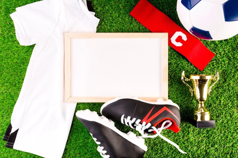 Cadre blanc posé sur une pelouse avec autour des objets rappelant le sport : t-shirt de sport, baskets à crampons, ballon de foot, trophée