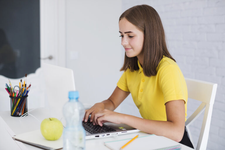 Jeune fille assise à son bureau travaillant sur un ordinateur portable. Sur le bureau sont posés un carnet, une bouteille d'eau, une pomme et un pot à crayons