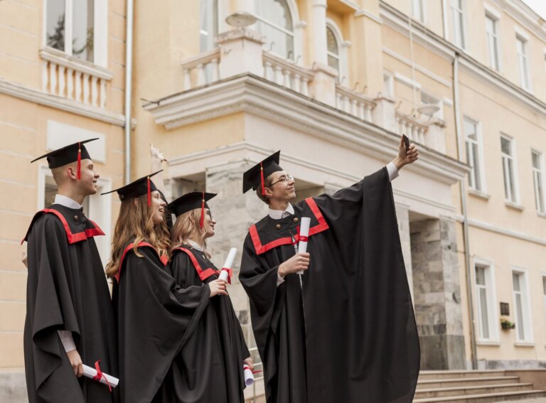 Groupe de 4 étudiant(e)s portant leur robe et couvre-chef noirs de diplômé(e)s, tenant leur diplôme entre les mains et prenant un selfie ensemble devant le bâtiment de leur école