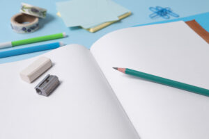 Cahier à pages blanches ouvert, plusieurs crayon posé sur et à côté du cahier, ainsi qu'une gomme et un taille-crayon
