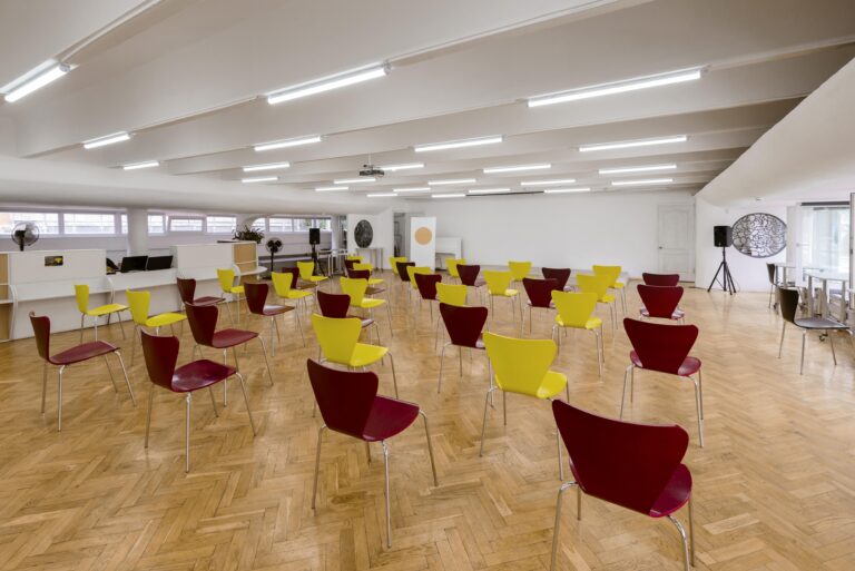 Photo à l'intérieur d'une salle quasiment vide au murs blancs avec des néons au plafond, le sol est en parquet clair. Dans la salle sont arrangées une trentaine de chaises bordeau et jaunes, toutes tournées vers le même mur
