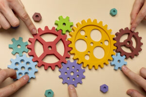 Des rouages en bois, de différentes tailles et dans des couleurs très vives, sont posés sur une table beige, cinq mains de personnes différentes semblent assembler les rouages entre eux