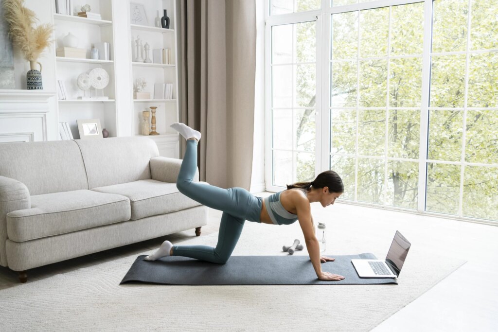 Femme dans son salon faisant du yoga sur un tapis de sport, un ordinateur portable est posé dessus, en arrière-plan il y a un canapé blanc et une bibliothèque blanche