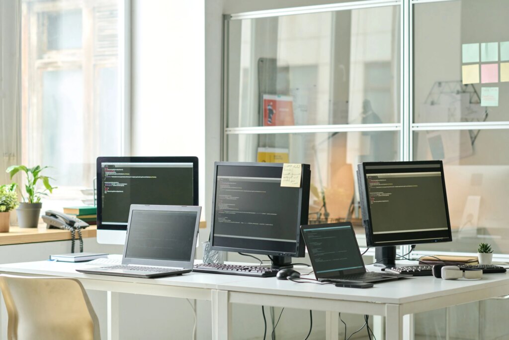 Vue d'un bureau dans un open space : au milieu une grande table avec 3 écrans et 2 ordinateurs portables affichant des lignes de code, en arrière-plan des post-its sont collés sur des parois vitrées