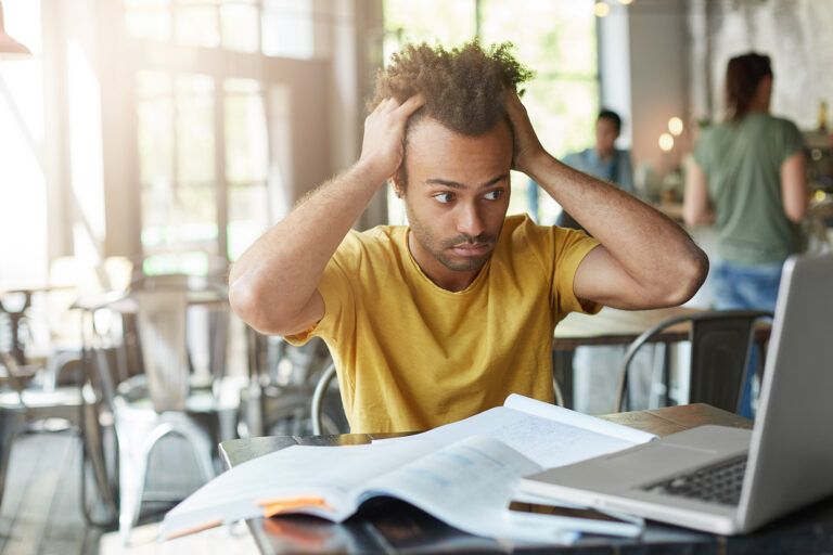 Trouver une alternance en urgence - Blog - étudiant assis dans un café se passant les mains dans les cheveux, en regardant son ordinateur d'un air stressé, des documents éparpillés sur la table devant lui