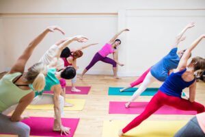 BPJEPS APT - Blog - Cours de sport dans une salle blanche avec du parquet, les participant(e)s font du yoga sur des tapis de gym colorés