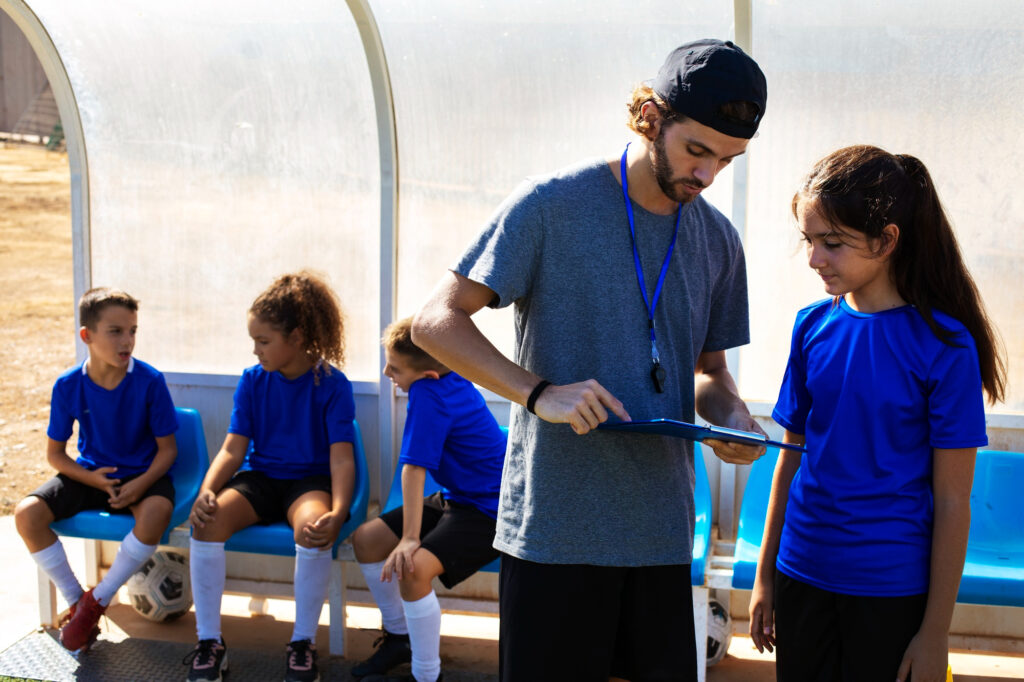 Jeune entraîneur de football masculin, casquette à l'envers et sifflet autour du cou, explique une tactique sur un clipboard à une joueuse adolescente en maillot bleu. Derrière lui, plusieurs enfants en uniformes bleus sont assis sur le banc de touche sous un abri transparent, avec des ballons de foot au sol.