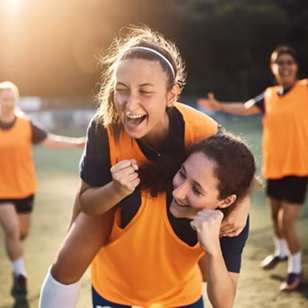 Deux joueuses de football souriantes et complices, l'une porte l'autre sur le dos pendant une célébration joyeuse sur le terrain