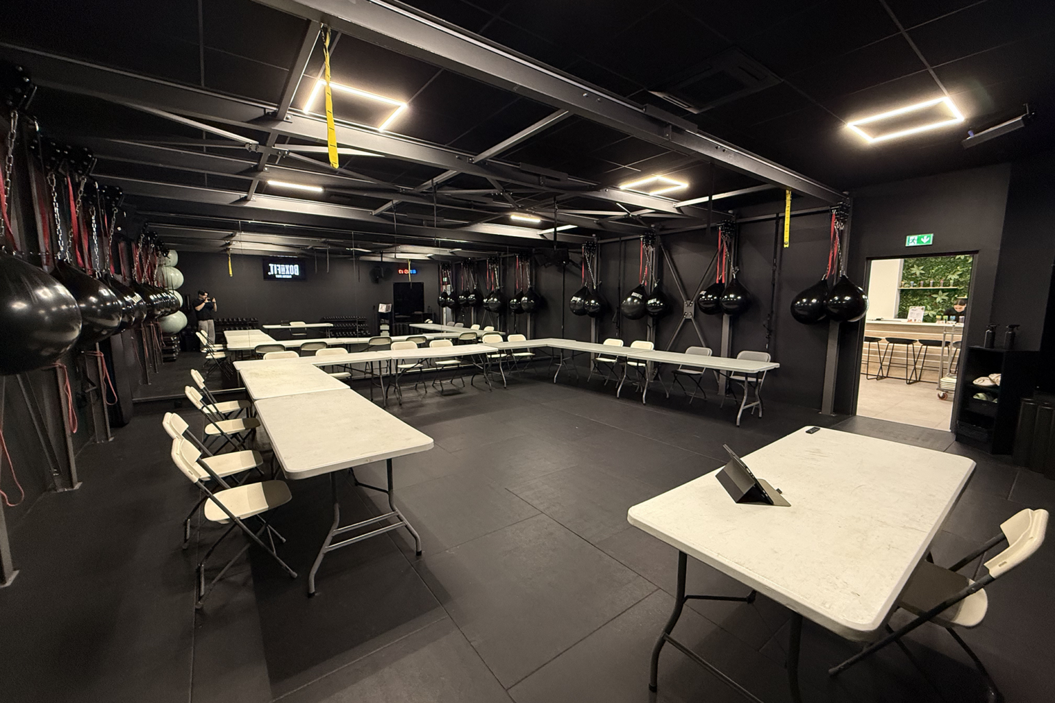 Salle de Boxe&Fit plongée dans une ambiance sombre avec éclairage LED, tables pliantes blanches disposées en U autour d'un grand espace central en tapis noir, sacs de frappe suspendus, haltères, ballons et horloge numérique visible au fond.