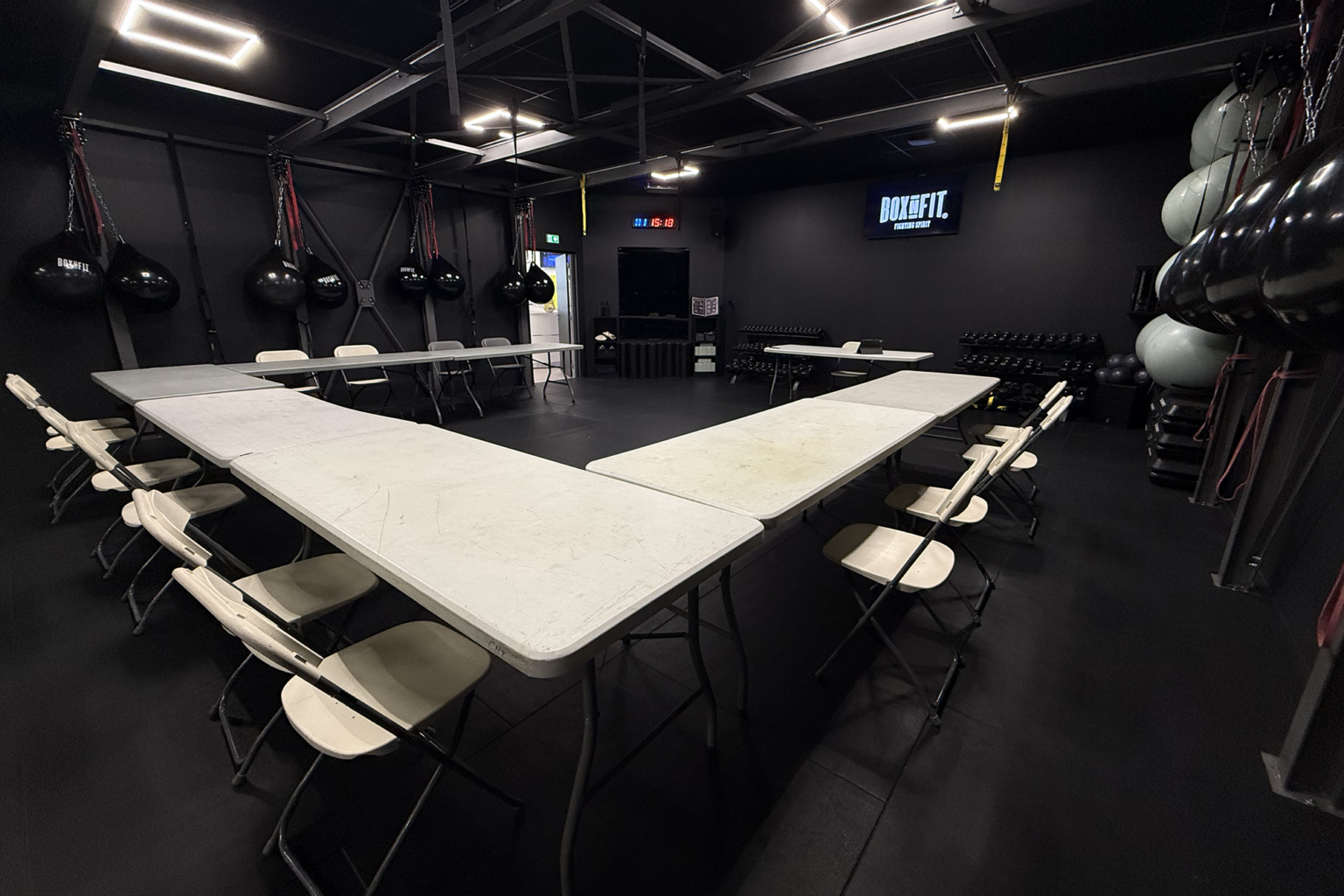 Salle de Boxe&Fit plongée dans une ambiance sombre avec éclairage LED, tables pliantes blanches disposées en U autour d'un grand espace central en tapis noir, sacs de frappe suspendus, haltères, ballons et horloge numérique visible au fond.