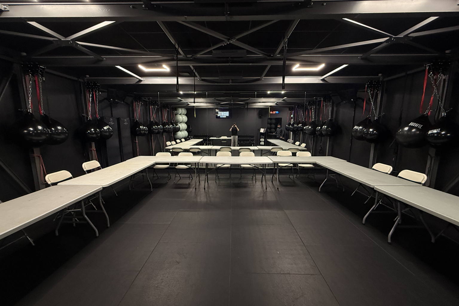 Salle de Boxe&Fit plongée dans une ambiance sombre avec éclairage LED, tables pliantes blanches disposées en U autour d'un grand espace central en tapis noir, sacs de frappe suspendus, haltères, ballons et horloge numérique visible au fond.