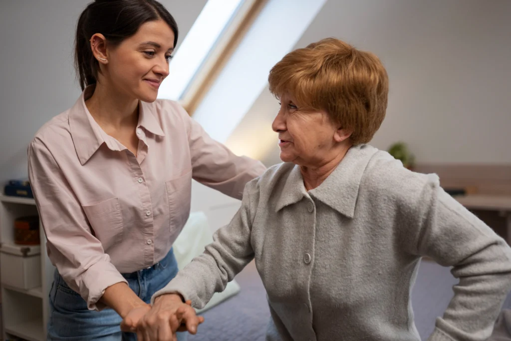Jeune femme aidante en chemise rose pâle soutient et accompagne avec douceur une dame âgée aux cheveux roux courts en gilet gris, toutes deux se tenant les mains et se regardant avec un sourire bienveillant dans une pièce lumineuse sous une grande fenêtre de toit.