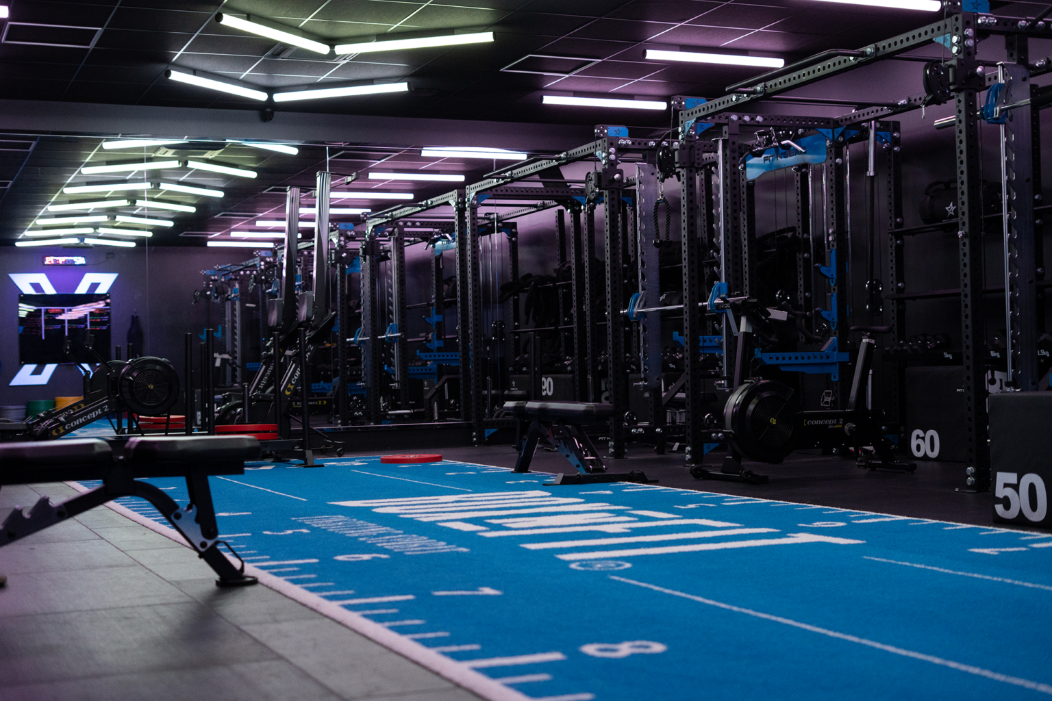 Salle de musculation moderne et sombre avec éclairage LED violet et blanc, grands miroirs couvrant un mur, cages à squat et racks noirs chargés de barres et poids, bancs, haltères, rameurs Concept2, sol bleu marqué 'BOXFIT' et zones numérotées pour les exercices.