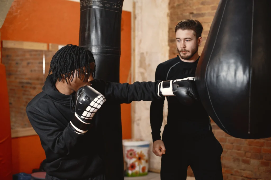 Boxeur aux dreadlocks en sweat noir à capuche frappe un sac de boxe lourd avec un direct du bras droit, gants noirs et blancs, tandis qu’un coach barbu en tenue noire moulante observe attentivement à ses côtés dans une salle aux murs de briques apparentes et sol en béton.