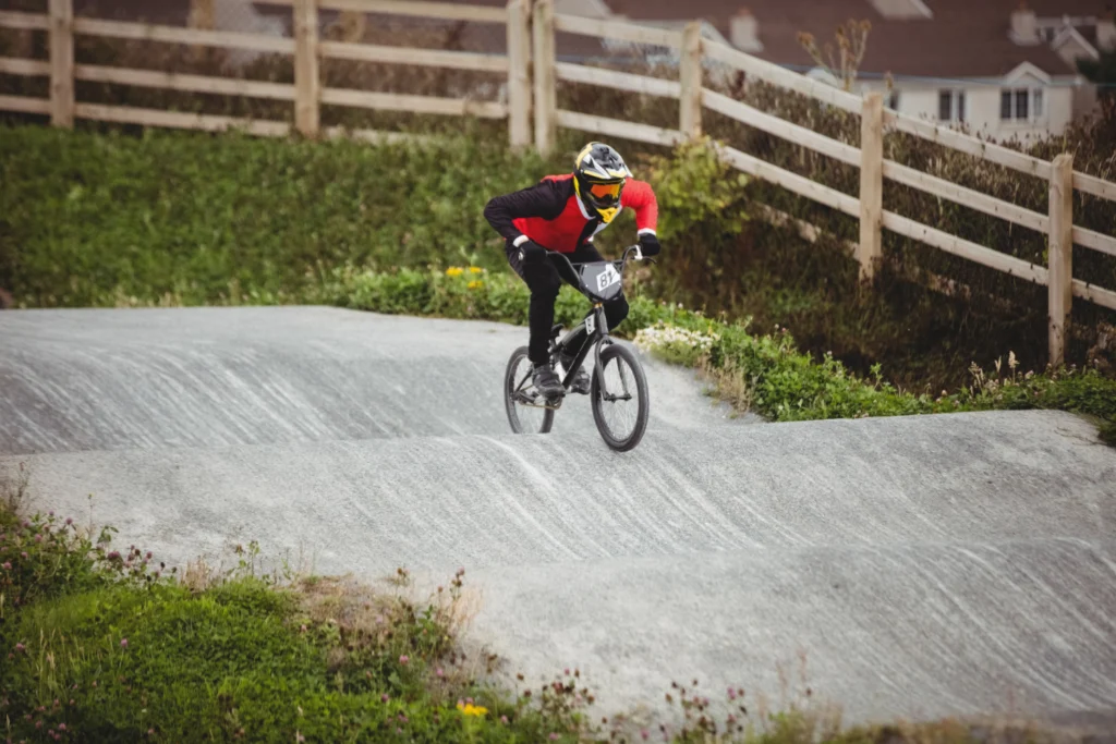 Jeune rider de BMX en combinaison rouge et noire, casque jaune et noir, lunettes orange, en plein saut sur une bosse d’un pumptrack extérieur en béton gris, penché en avant sur son vélo noir avec plaque 81, entouré de talus herbeux, barrières en bois et maisons en arrière-plan.