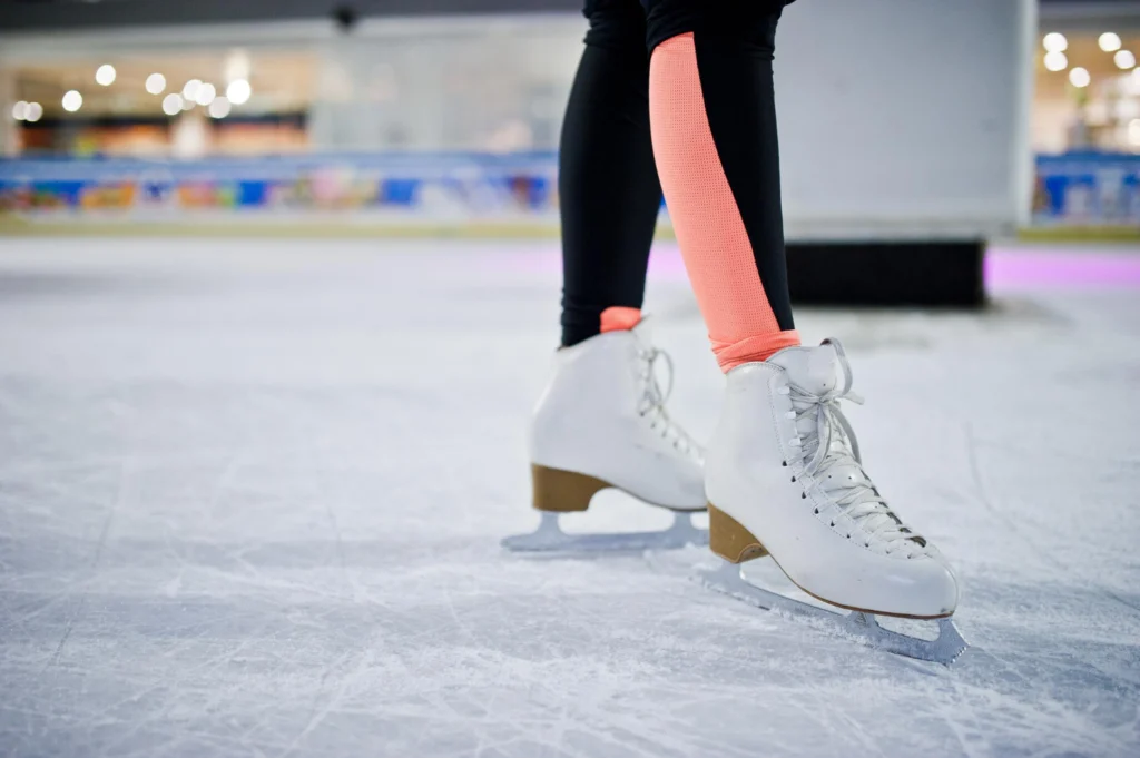 Gros plan sur les patins à glace blancs d'une patineuse, lacets bien attachés, posés sur la glace rayée d'une patinoire intérieure, avec des leggings noirs et bandes roses flashy sur les mollets en arrière-plan flou.